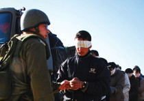 An IDF soldier stands next to a blindfolded Palestinian prisoner