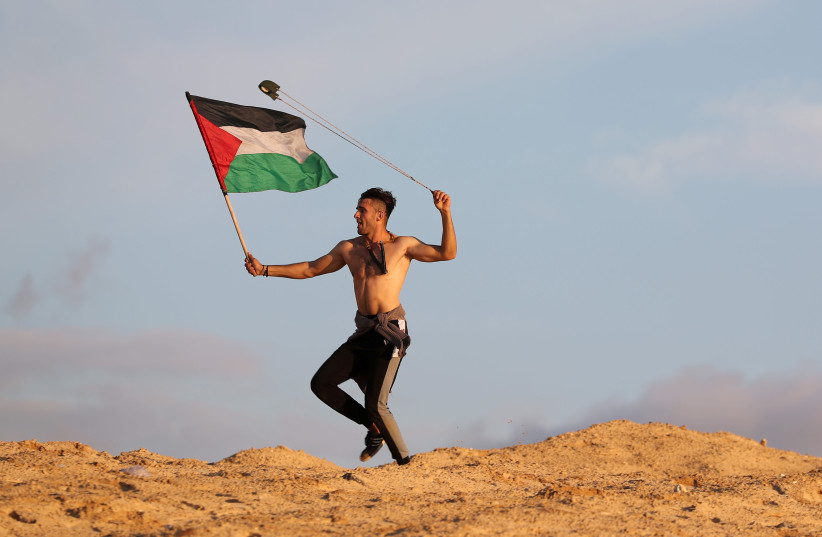 A demonstrator holding a Palestinian flag uses a sling to hurl stones at Israeli troops during a protest calling for lifting the Israeli blockade on Gaza, at the beachfront border with Israel, in the northern Gaza Strip November 5, 2018 (photo credit: MOHAMMED SALEM/REUTERS)