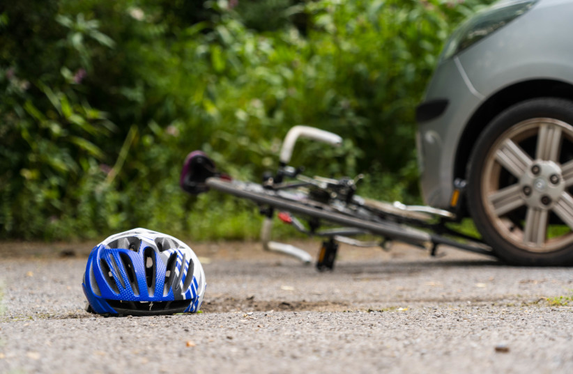  Photo of a helmet lying on ground with bicycle trapped underneath a car in the background during a road traffic accident involving a bike crash. (illustrative). (photo credit: Wikimedia Commons)