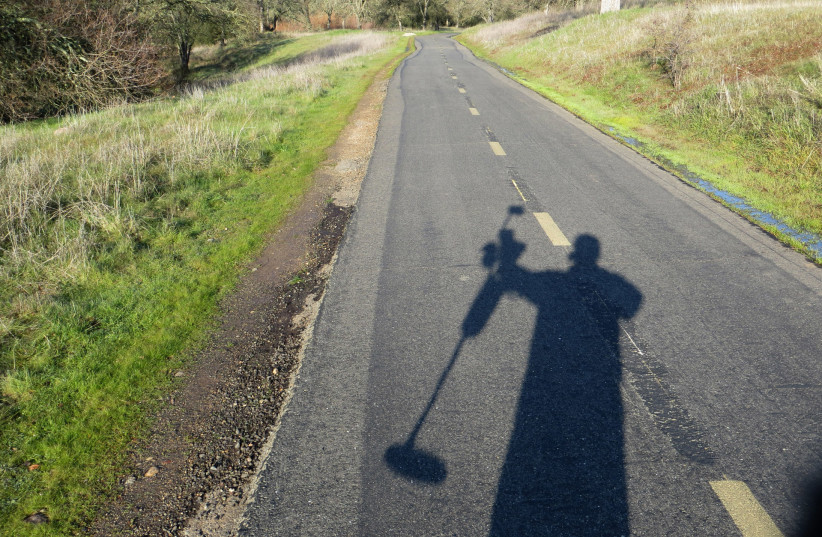  The shadow of a man holding a metal detector is visible on the road near woods.  (photo credit: PUBLICDOMAINPICTURES.NET)