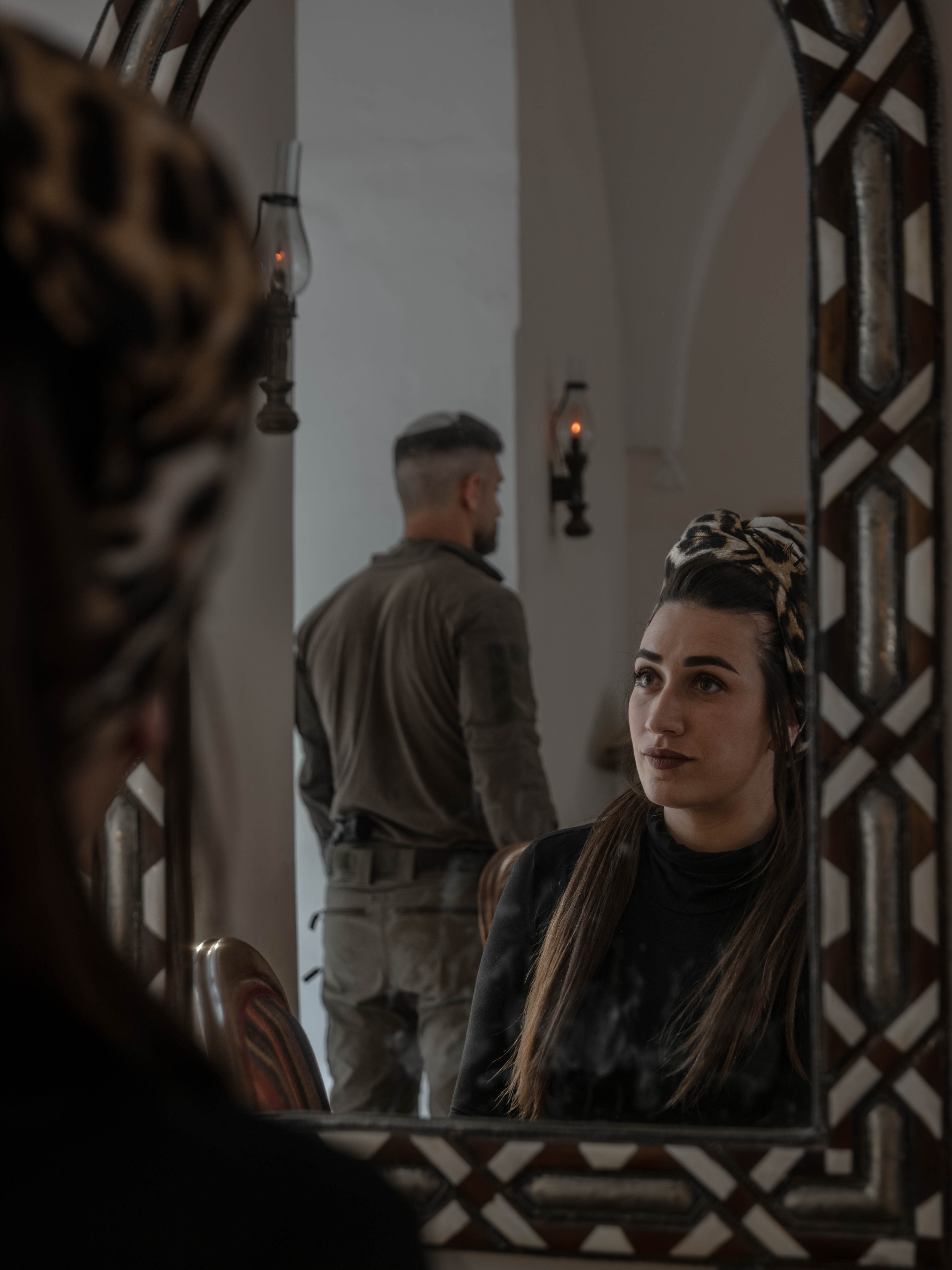 ELIANA WATCHES her husband step back into uniform, her reflection caught in the mirror – a quiet moment that captures the emotional split between holding a household together and living with the constant uncertainty of war.