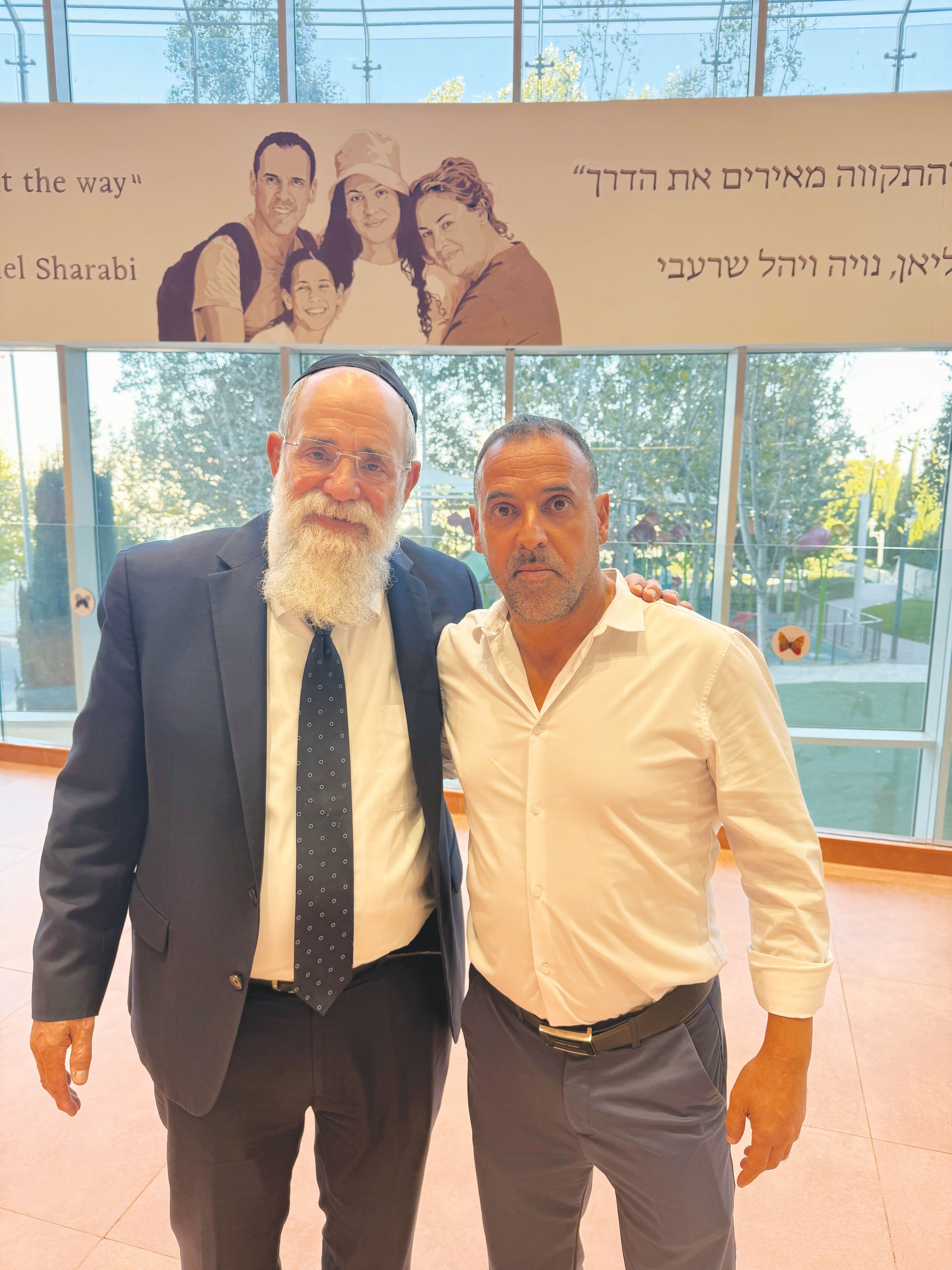 KALMAN SAMUELS (L) founder-president of Shalva, with Eli Sharabi, next to the drawing of Eli’s family, in the main lobby of Shalva’s headquarters in Jerusalem. =