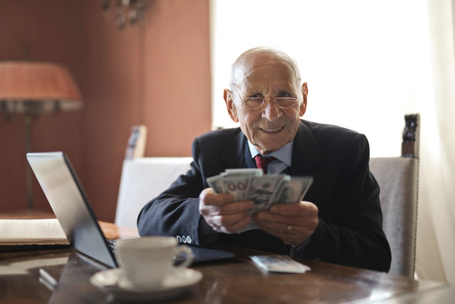 A senior businessman holding money in hands while sitting at table near laptop. (Illustrative) (credit: Andrea Piacquadio/Pexels)