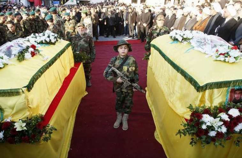 The son of Hezbollah operative Abbas Hijazi, who died in an airstrike in Quneitra, carries a toy weapon as he stands between his father's (R) and grandfather's (L) coffins in Ghaziyeh village (photo credit: REUTERS)