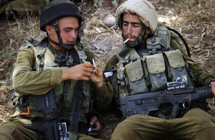 IDF soldiers share cigarettes while resting in the shade (credit: FINBARR O'REILLY / REUTERS)