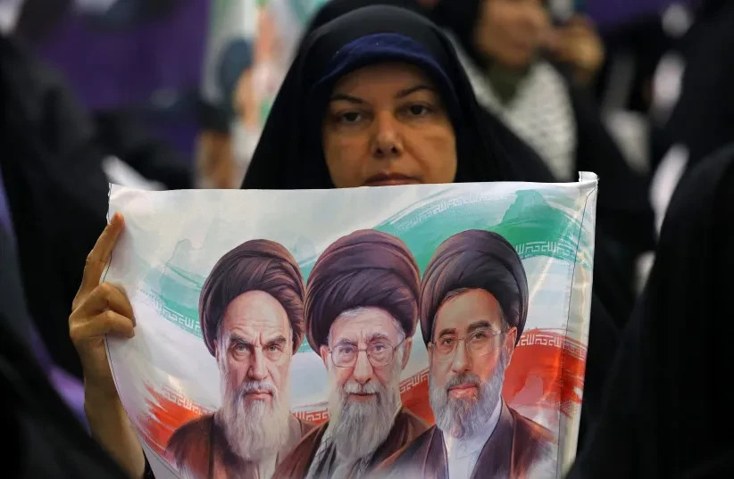 Una mujer iraní sostiene una pancarta con las imágenes de los tres líderes supremos iraníes superpuestas sobre la bandera de la República Islámica en una manifestación contra Israel y Estados Unidos celebrada en Teherán el 24 de mayo de 2026; imagen ilustrativa.  (credit: Atta Kenare/AFP via Getty Images)