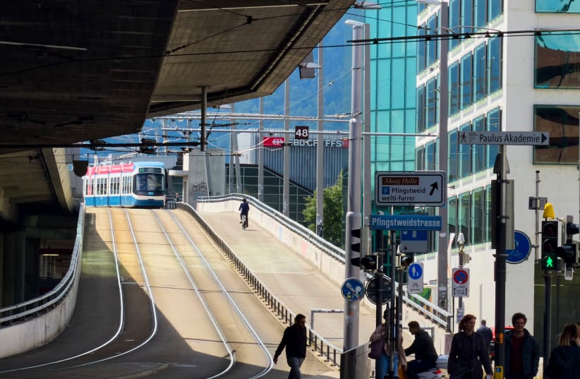 Crowd of pedestrians and commuters cross a busy urban intersection by a tram ramp and underpass at Hardbrücke, framed by modern office buildings and street signs on May 19, 2026 in Zurich, Switzerland.
