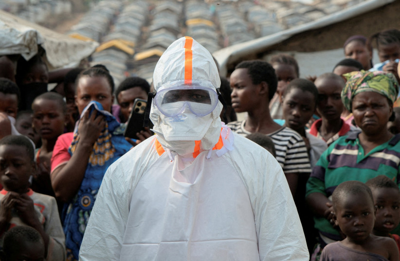 A health worker in personal protective equipment (PPE) stands near displaced people waiting for the burial of suspected Ebola victims at the Kigonze displaced persons camp, one month after an outbreak was declared, in Bunia, eastern Democratic Republic of Congo, June 18, 2026.