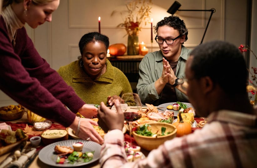 A group of young adults is seen sitting around a table sharing a meal, eating and talking.