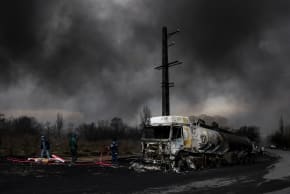 People stand near a destroyed vehicle as smoke rises after a reported strike on Shahran fuel tanks, in Tehran, Iran, March 8, 2026