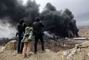 People walk as smoke rises in the background after a reported strike on Shahran fuel tanks, in Tehran, Iran, March 8, 2026