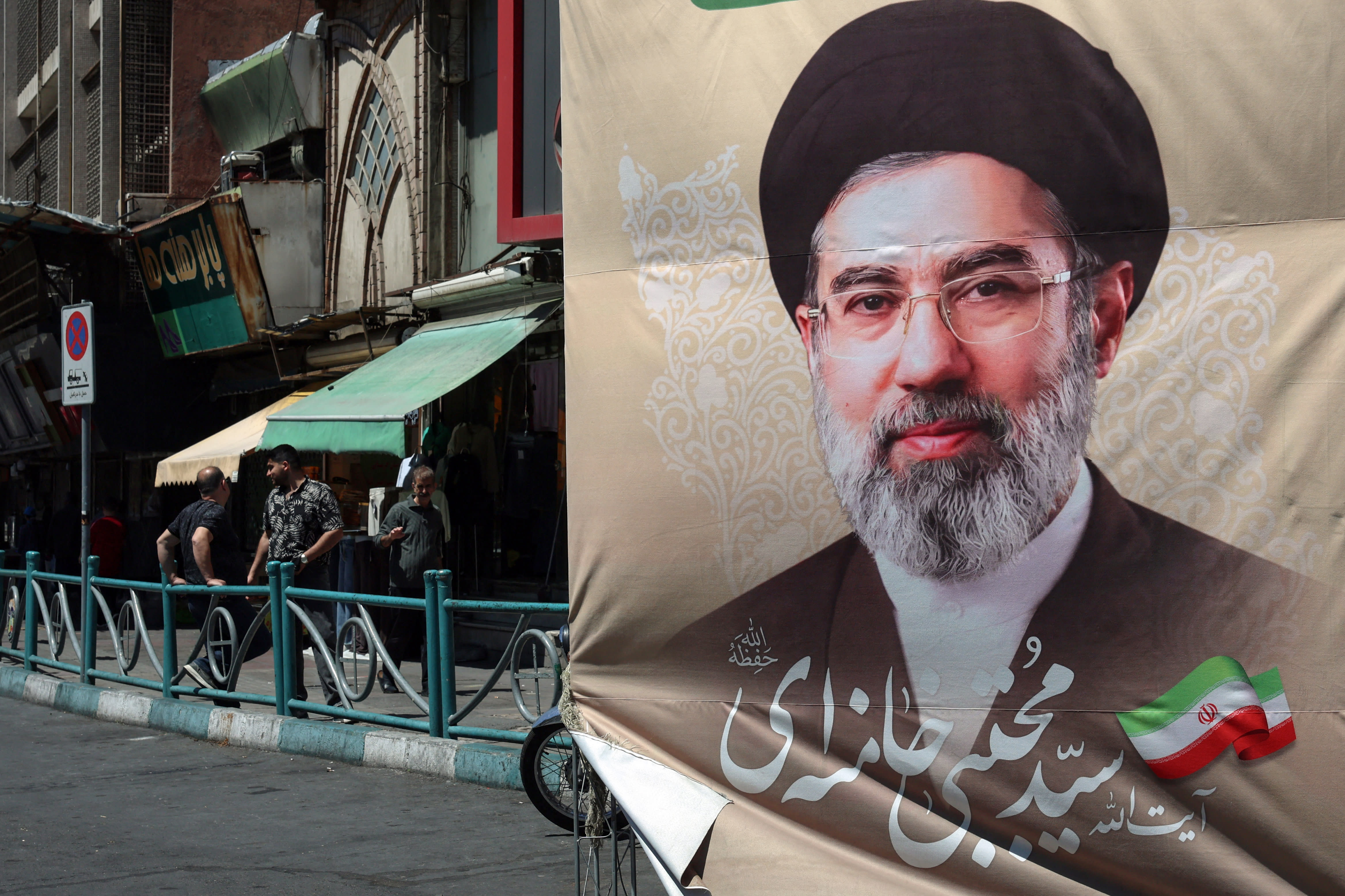 Iranians stand on a pavement along a street next to a billboard depicting Iran