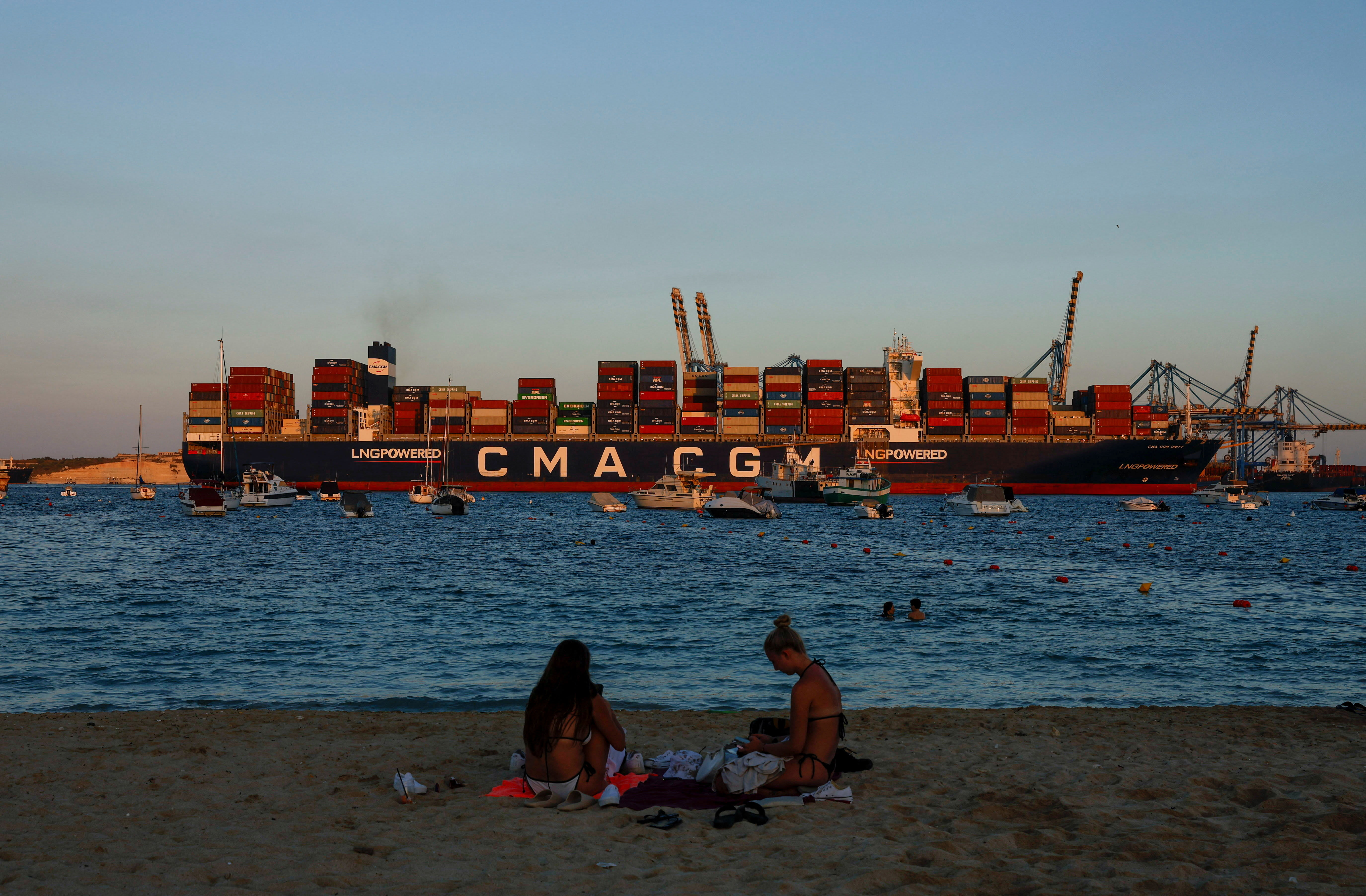 People relax on a beach as the container ship CMA CGM UNITY arrives at Malta Freeport, in Birzebbuga, Malta September 18, 2025. (photo credit: DARRIN ZAMMIT LUPI/REUTERS)