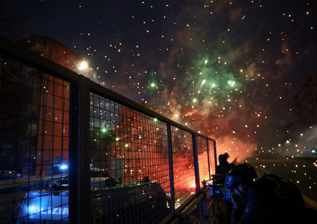 A person uses a camera while standing behind a fence next to a police vehicle, with fireworks exploding as demonstrators use them against police officers while trying to block a road leading to the Santa Giulia Arena, on the day of a protest against the environmental, economic and social impact. 