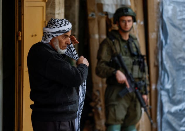 A Palestinian man puts on a keffiyeh as an Israeli soldier stands guard in the old city in Hebron in the West Bank, February 9, 2026.
