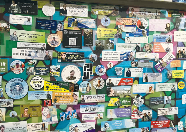 A station wall layered with portraits, stickers, and memorial notes – an everyday map of loss, kept in public view long after the headlines moved on. 