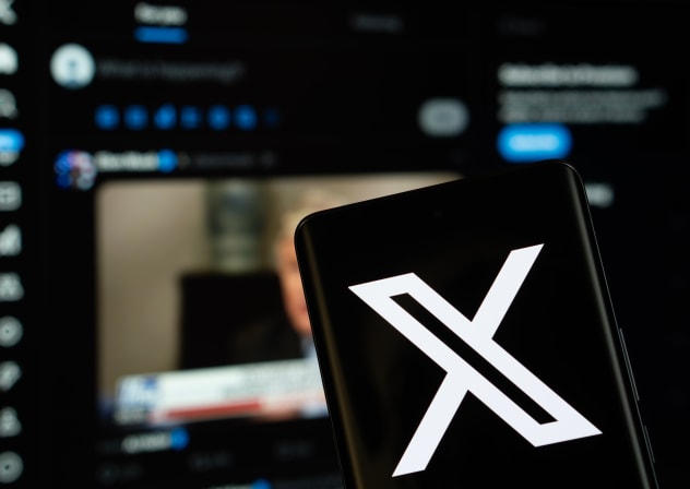 X (twitter) logo is seen on a smartphone, with the bluesky website visible on a computer screen in the background.