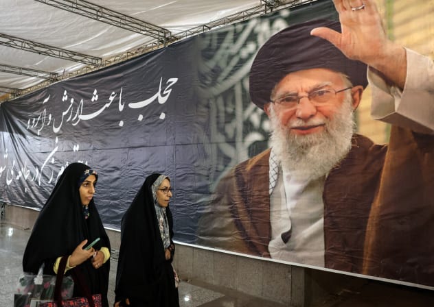 Iranian women walk past a banner bearing a portrait of Iran's supreme leader Ayatollah Ali Khamenei during the Iranian-Islamic fashion design fair at the Imam Khomeini Grand Mosque in Tehran on February 22, 2026.
