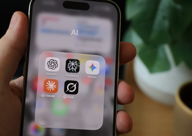 A person holding a smartphone displaying an AI folder with icons for ChatGPT, Perplexity, Gemini, Claude, and Grok among a backdrop of greenery.