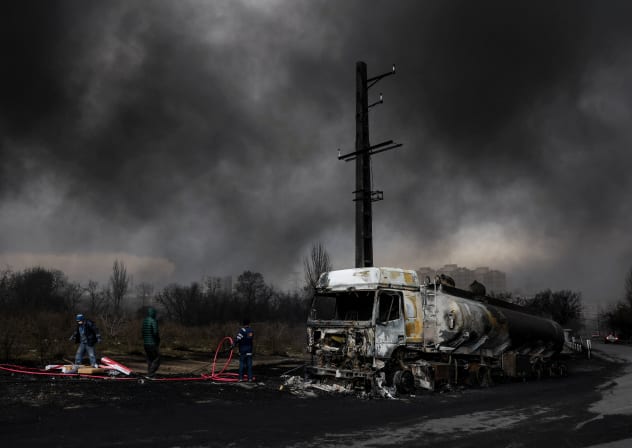 People stand near a destroyed vehicle as smoke rises after a reported strike on Shahran fuel tanks, in Tehran, Iran, March 8, 2026