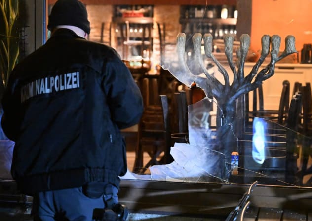 A detective stands with a flashlight in front of the smashed window of an Israeli restaurant in Munich, Germany, April 10, 2026. 
