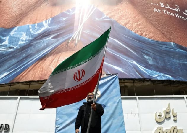 Un hombre sostiene una bandera iraní junto a una valla publicitaria contra Estados Unidos en la que aparecen el presidente estadounidense Donald Trump y el estrecho de Ormuz, en Teherán (Irán), el 2 de mayo de 2026. 