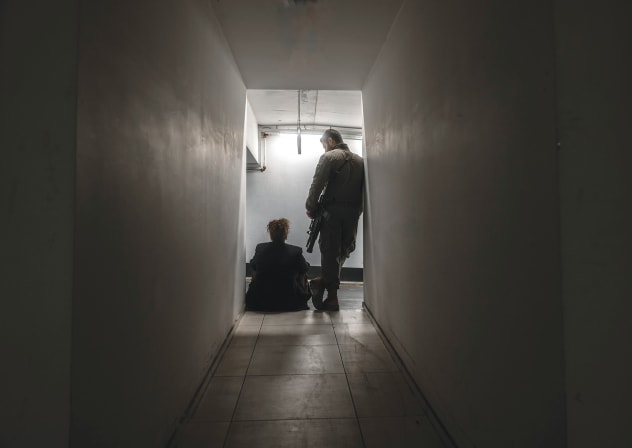 Neora and E. moved to Ramat Gan together two years ago and are looking forward to starting a family. Here, Neora sits in a narrow hallway as her partner stands above her in uniform, the weight of absence lingering even in proximity.