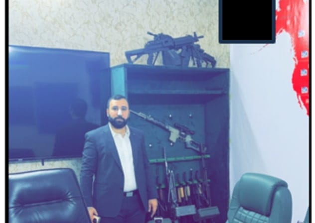 MOHAMMAD BAQER Saad Dawood Al-Saadi standing in front of an array of machineguns in a conference room.