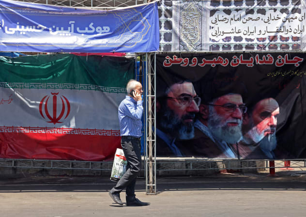 A man walks past Iran’s national flag and a banner bearing the images of the late founder of the Islamic Revolution, supreme leader Ayatollah Ruhollah Khomeini (R), the late Iranian supreme leader Ali Khamenei and his son, the current supreme leader Mojtaba Khamenei (L), in Tehran, June 10.