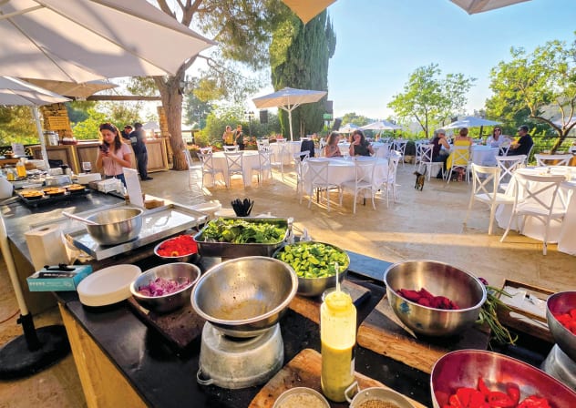 A vibrant, sun-drenched view of the open-air terrace at the Logos Hotel, where an expansive farm-to-table prep station loaded with fresh, raw ingredients sets the stage for Luiza’s monthly ‘Meshak Shuk’ culinary festival.