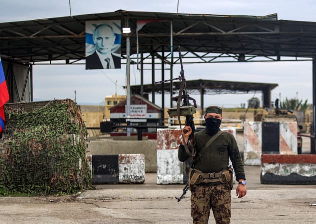 A Syrian rebel fighter (C) looks on as he stands at an inspection checkpoint for incoming vehicles before a Russian flag and Russian soldiers behind manning the entrance of the Russian-leased Syrian military base of Hmeimim in Latakia province in western Syria on December 29, 2024.