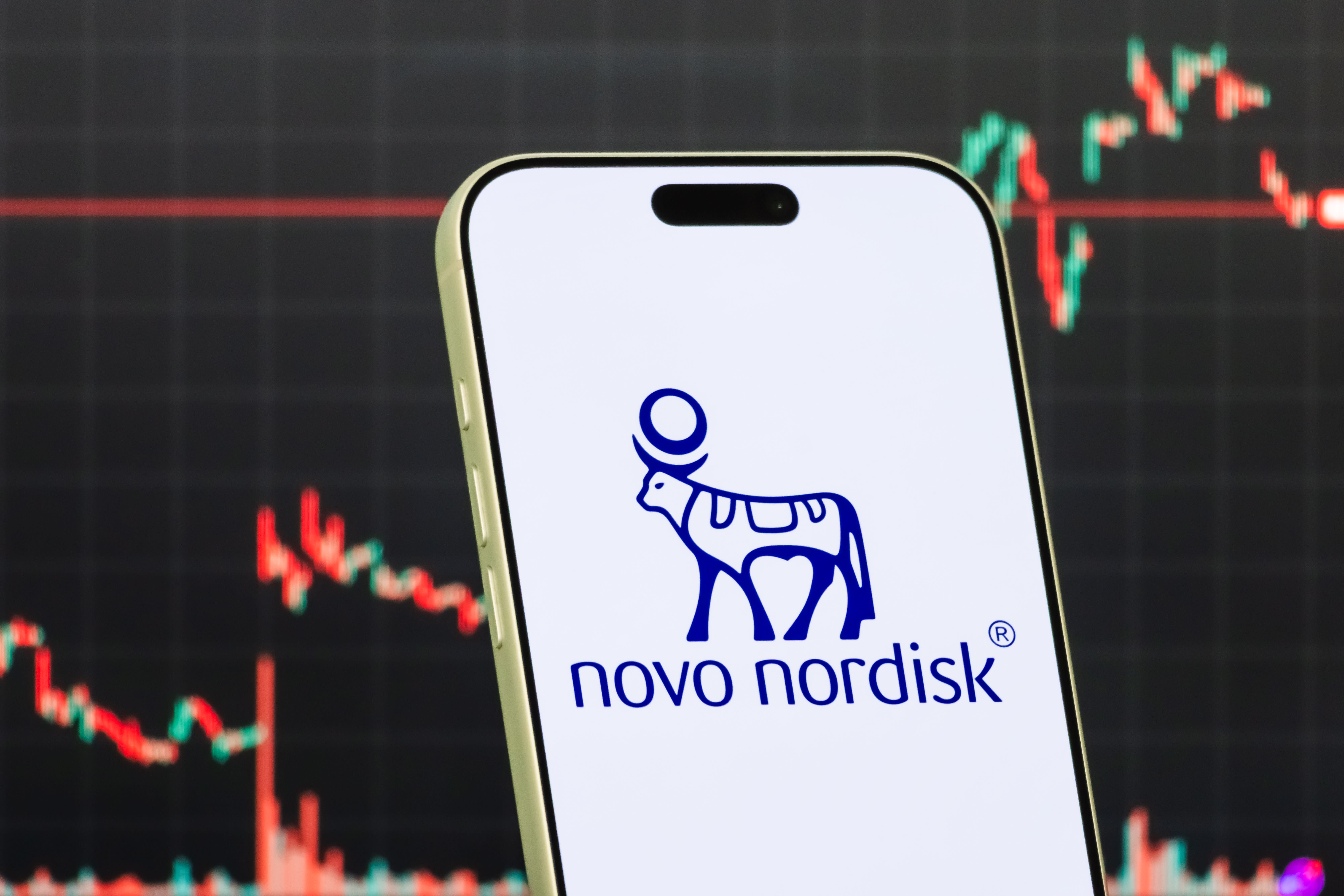 In this photo illustration, a smartphone displays the logo of Novo Nordisk (NYSE: NVO), a Denmark-based pharmaceutical company specializing in diabetes care, obesity treatment, in front of a screen showing the company