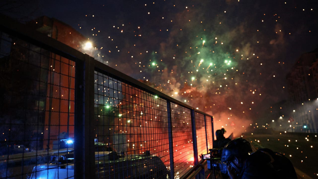 A person uses a camera while standing behind a fence next to a police vehicle, with fireworks exploding as demonstrators use them against police officers while trying to block a road leading to the Santa Giulia Arena, on the day of a protest against the environmental, economic and social impact. 
