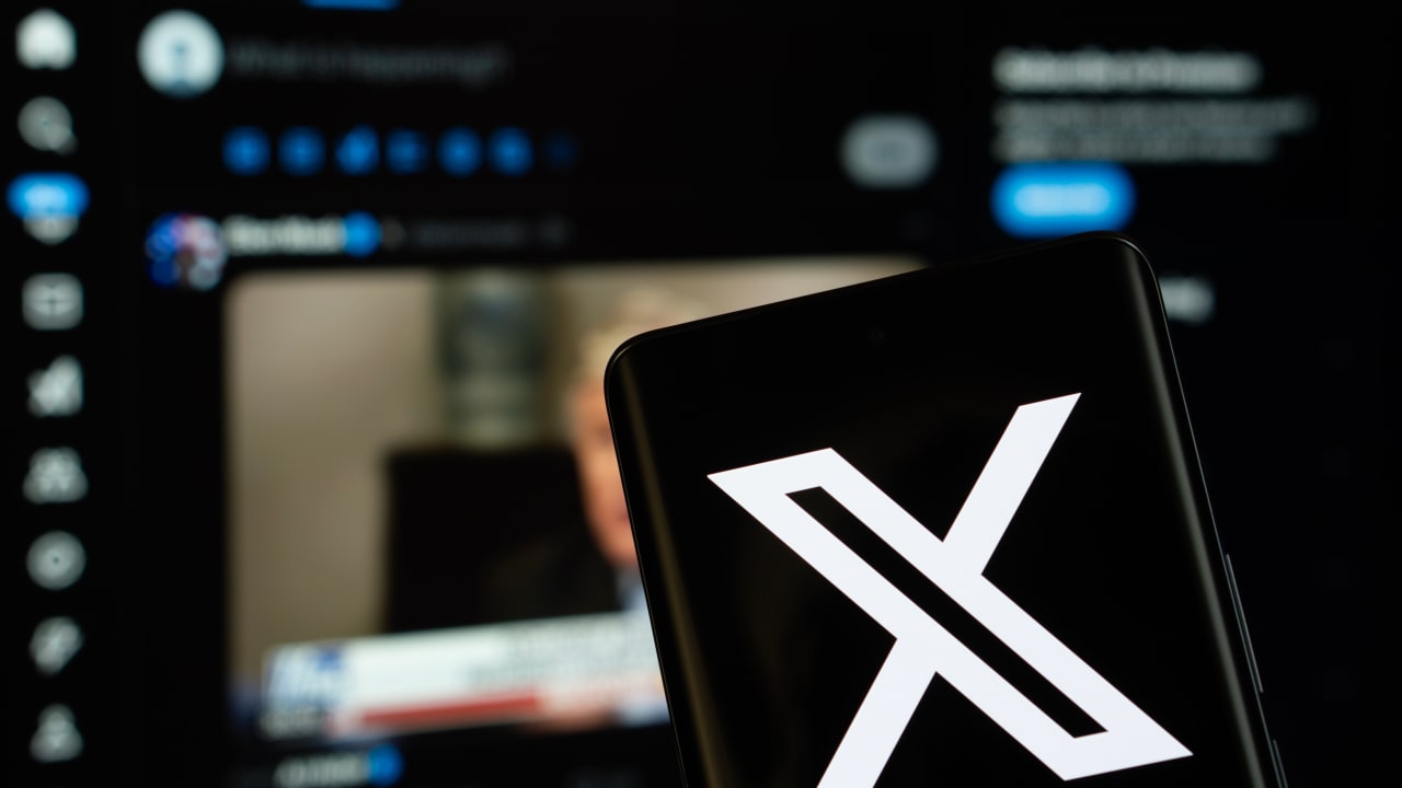 X (twitter) logo is seen on a smartphone, with the bluesky website visible on a computer screen in the background.