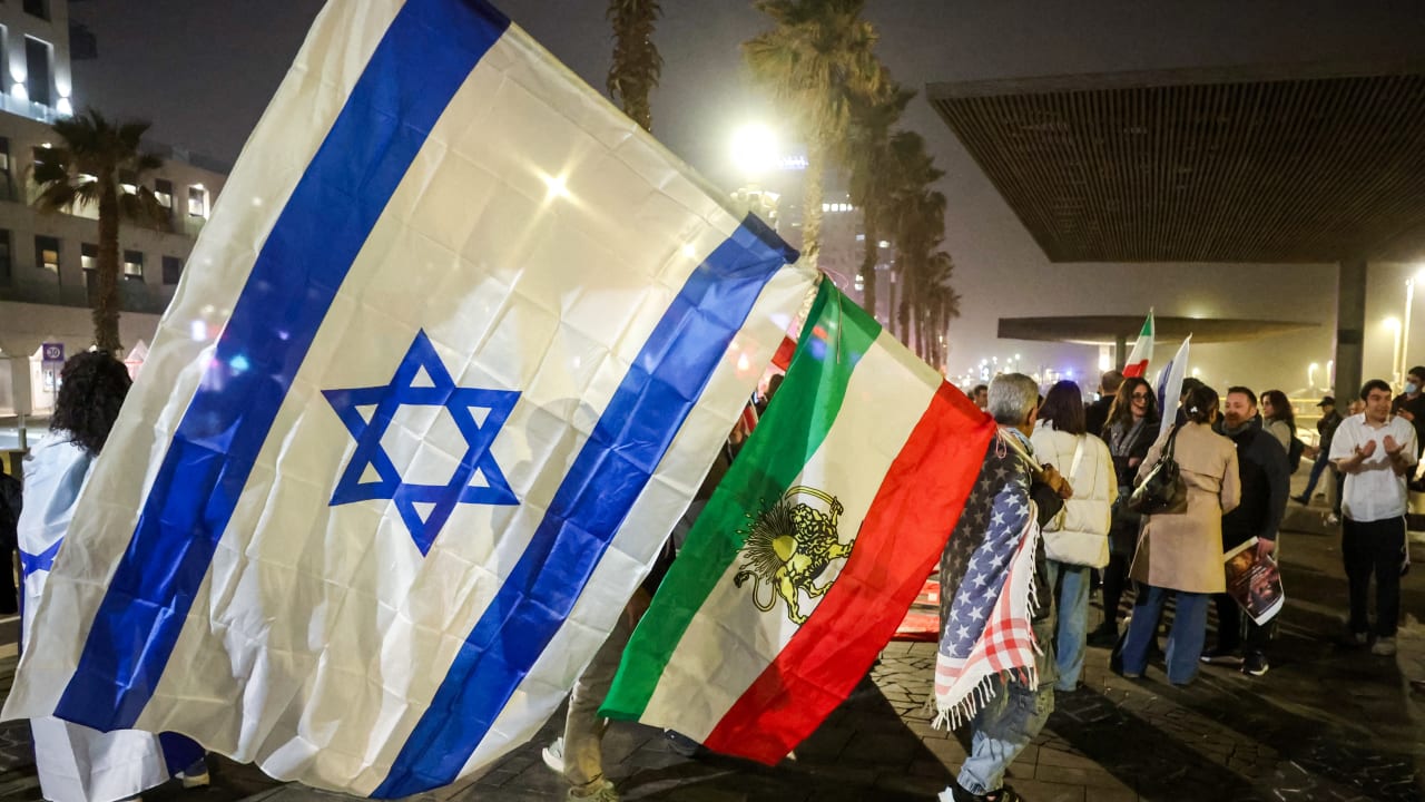 A man draped in a US flag holds stacked Israeli and Royal Iranian flags during a demonstration in solidarity with the Iranian protest movement outside the US Embassy branch office in Tel Aviv on February 14, 2026. 