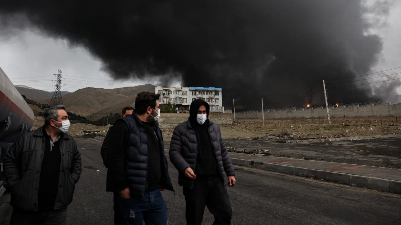 People walk as smoke rises in the background after a reported strike on Shahran fuel tanks, in Tehran, Iran, March 8, 2026