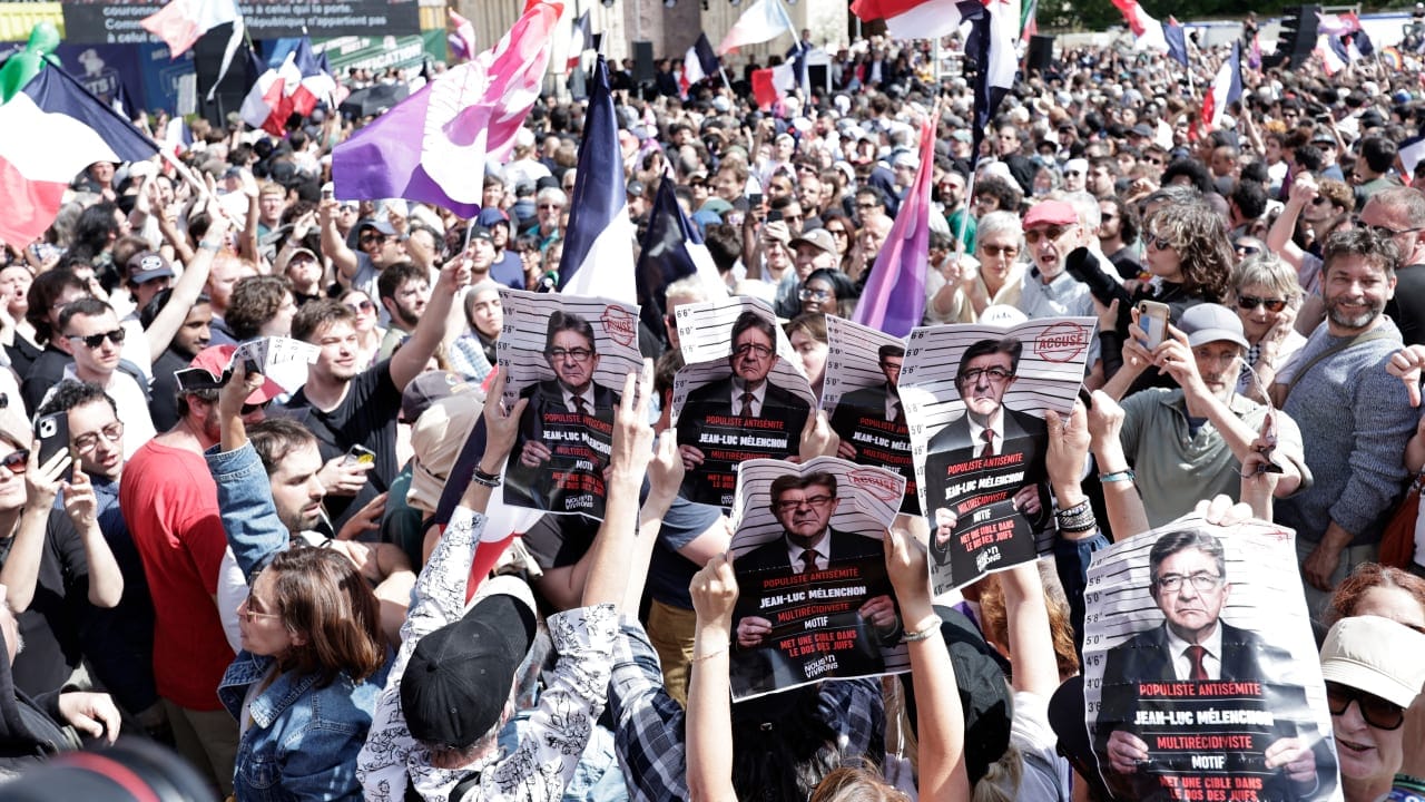 Protesters hold signs depitcting Jean-Luc Melenchon as in a mugshot and reading "Antisemite Populist, Jean-Luc Melenchon, Repeat offender, Puts a target in the back of Jews." 