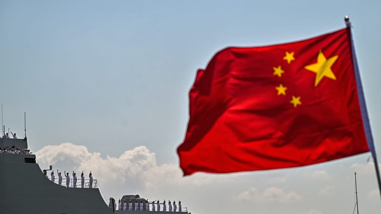 Crew members of Chinese People's Liberation Army (PLA) navy ship Qi Jiguang wave as their ship prepares to dock past a flag of China at Tanjung Perak Port in Surabaya on August 2, 2026, in response to an invitation from the Indonesian Navy. 