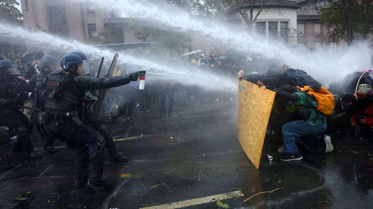 Protesters take cover behind a plywood board as French riot police and gendarmes use a water canon amid clashes during an authorised demonstration organised by labor unions in Paris, October 24, 2026.
