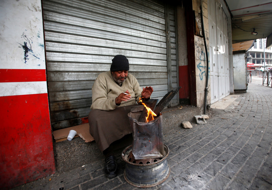 A Palestinian warms himself outside a row of closed shops in Hebron as part of a general Palestinian strike against US VP Pence's visit, January 2018 (Credit: Mussa Qawasma/ Reuters)