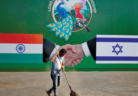 A municipal worker cleans the street in front of a bilboard displaying Indian and Israeli flags for 