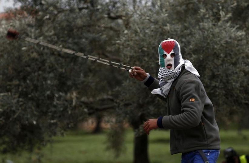 A Palestinian protester wearing a mask featuring the Palestinian flag, uses a slingshot to hurl stones towards Israeli troops in Bil'in (photo credit: REUTERS)