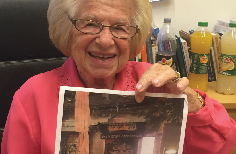 An icon in spraypaint: Dr. Ruth holds up a photo of a storefront in Mahaneh Yehuda that has been decorated with her smiling face (one of the many done by Solomon Souza of influential/historical Jewish figures). (photo credit: ARIEL DOMINIQUE HENDELMAN)