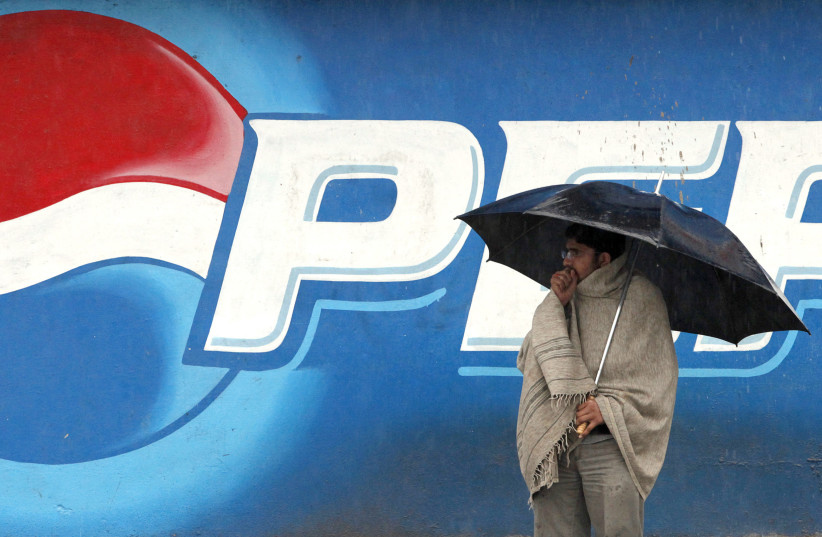 A man stands next to a Pepsi advertisement while using an umbrella in the rain, in Rawalpindi (photo credit: FAISAL MAHMOOD/REUTERS)