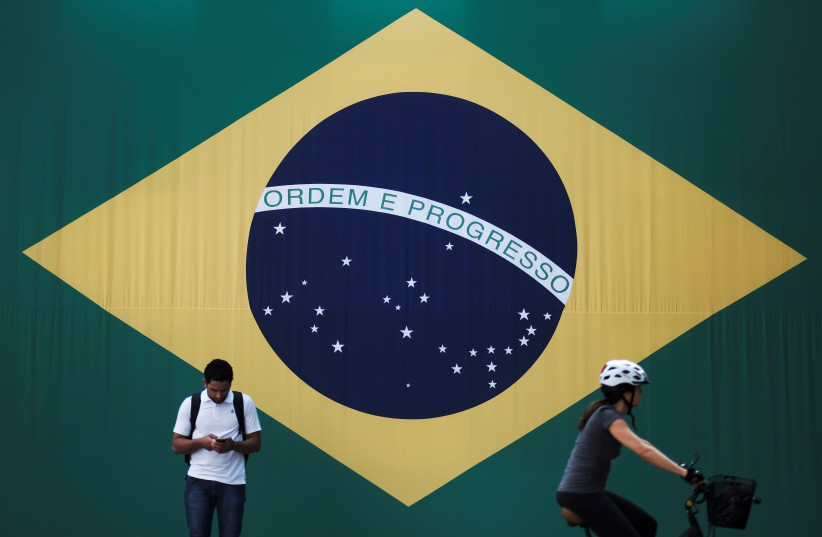 A man checks his mobile phone as a woman riding a bike passes next to a big Brazilian flag in Sao Paulo, Brazil June 28, 2018. (credit: REUTERS/NACHO DOCE)