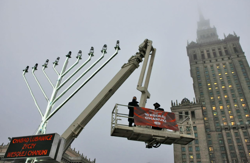 Warsaw Mayor Rafał Trzaskowsk and Chabad of Poland's Rabbi Shalom Ber Stambler are seen being lifted to the top of a large menorah as part of a Hanukkah celebration in front of the Palace of Culture. (photo credit: CHABAD WARSAW)
