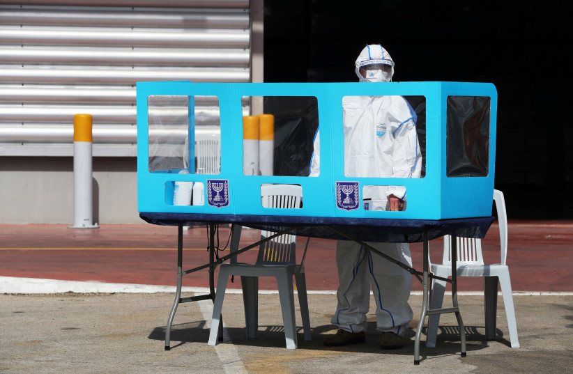 An employee wearing personal protective equipment stands behind a booth during a demonstration revealing preparations ahead of Israel’s upcoming election, at the Central Elections Committee’s logistics center in Shoham (photo credit: AMMAR AWAD / REUTERS)