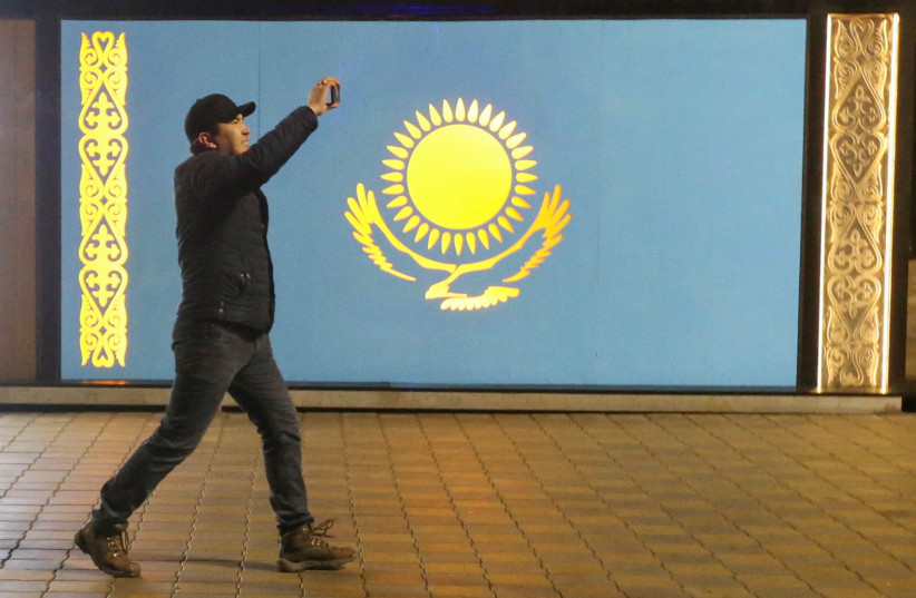  A man holds a mobile phone while walking past a board with a Kazakh state flag during a protest against LPG cost rise following authorities' decision to lift price caps on liquefied petroleum gas in Almaty, Kazakhstan January 5, 2022 (photo credit: REUTERS/PAVEL MIKHEYEV)