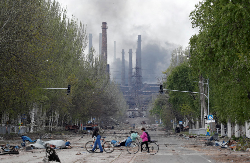  People walk their bikes across the street as smoke rises above a plant of Azovstal Iron and Steel Works during Ukraine-Russia conflict in the southern port city of Mariupol, Ukraine May 2, 2022.  (credit: REUTERS/ALEXANDER ERMOCHENKO)