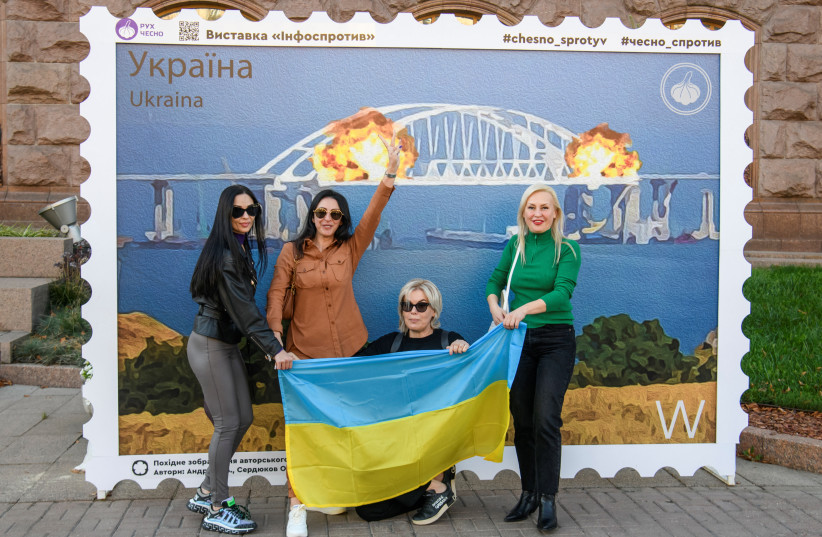  Women pose with the Ukrainian national flag next to an artwork depicting Kerch bridge on fire, amid Russia's attack on Ukraine, in central Kyiv, Ukraine October 8, 2022. (credit: REUTERS/VLADYSLAV MUSIIENKO)